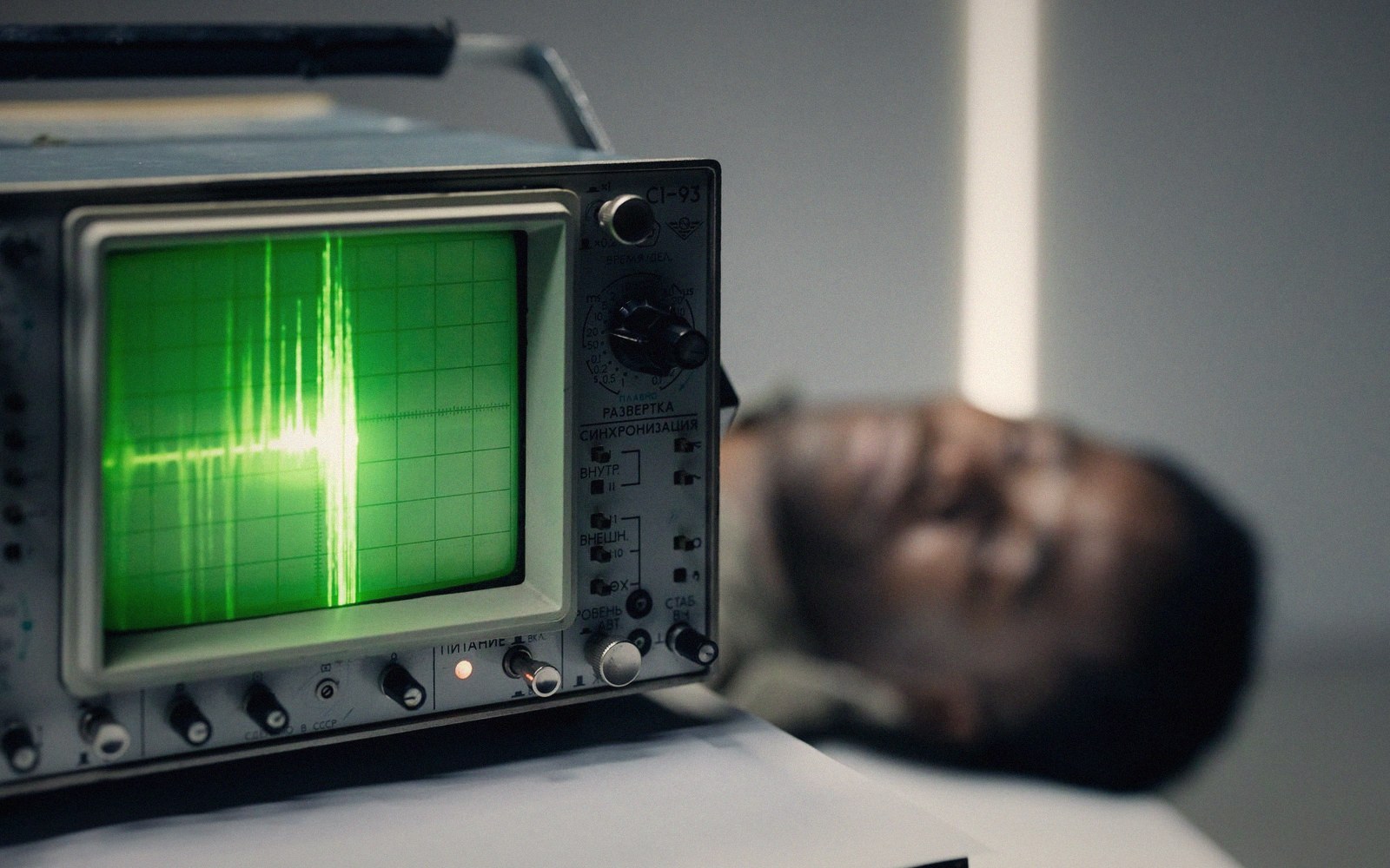 An oscilloscope displays a green waveform beside a person lying with eyes closed